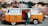 A colorful vintage van is parked in front of a graffiti-covered wall. The van is primarily orange and white, with 'PHOTO BUS' written on its side. Above it, a bright neon sign reads 'PHOTOS.' A man stands casually next to the van, wearing jeans, glasses, and a white t-shirt with an open jacket. The graffiti mural on the wall features a detailed, expressive face, contributing to the vibrant urban atmosphere.