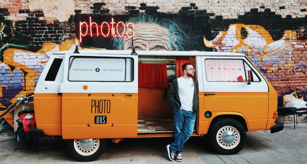 A colorful vintage van is parked in front of a graffiti-covered wall. The van is primarily orange and white, with 'PHOTO BUS' written on its side. Above it, a bright neon sign reads 'PHOTOS.' A man stands casually next to the van, wearing jeans, glasses, and a white t-shirt with an open jacket. The graffiti mural on the wall features a detailed, expressive face, contributing to the vibrant urban atmosphere.