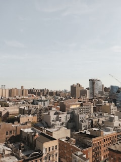 An urban skyline featuring a dense cluster of buildings with varied architectural styles, including modern high-rises and older brick structures. A construction crane is visible in the distance, and a pale blue sky stretches above the cityscape.