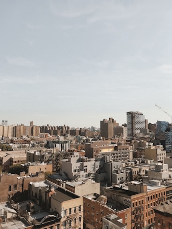 An urban skyline featuring a dense cluster of buildings with varied architectural styles, including modern high-rises and older brick structures. A construction crane is visible in the distance, and a pale blue sky stretches above the cityscape.