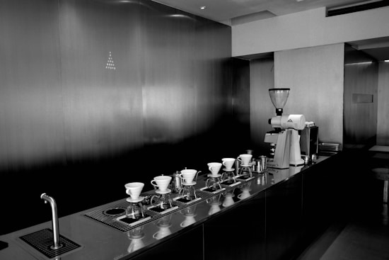 A sleek and modern coffee preparation area with multiple pour-over coffee drippers arranged in a row on a counter. There is a large coffee grinder on the right and a stainless steel faucet on the left. The environment is minimalistic, with a strong emphasis on clean lines and a monochromatic color palette.