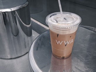 A cup of iced coffee with a straw in a transparent plastic cup is placed on a reflective surface. The cup has a logo or text on it. The background features metallic or chrome surfaces, giving a modern and sleek appearance.