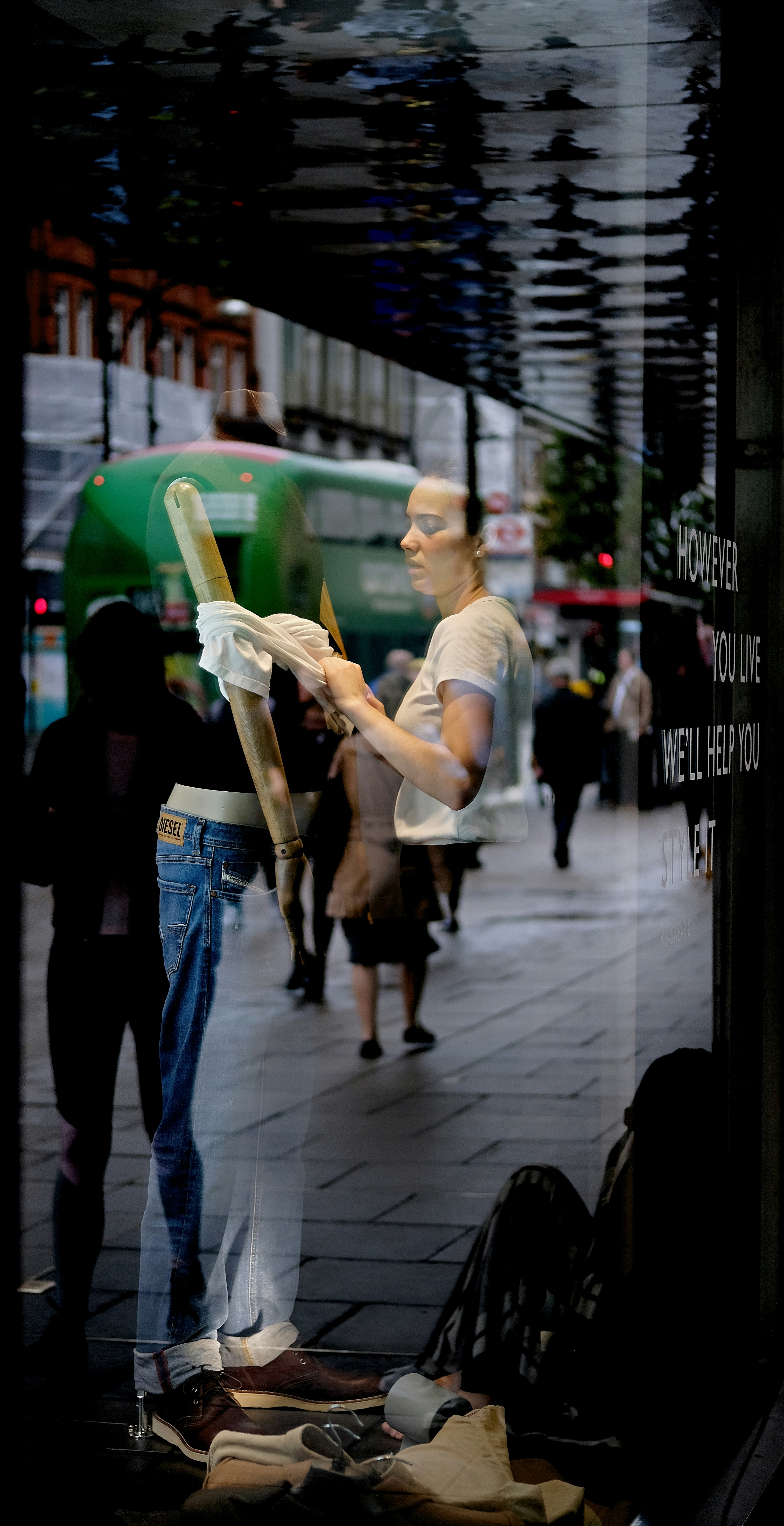 A person is seen cleaning a shop window from the inside, with reflections of a busy street outside. The person is wearing casual clothes and is holding a cloth. The display features jeans and brown shoes. Pedestrians and a green bus are visible in the reflection.