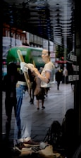A person is seen cleaning a shop window from the inside, with reflections of a busy street outside. The person is wearing casual clothes and is holding a cloth. The display features jeans and brown shoes. Pedestrians and a green bus are visible in the reflection.