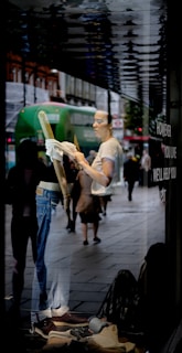 A person is seen cleaning a shop window from the inside, with reflections of a busy street outside. The person is wearing casual clothes and is holding a cloth. The display features jeans and brown shoes. Pedestrians and a green bus are visible in the reflection.