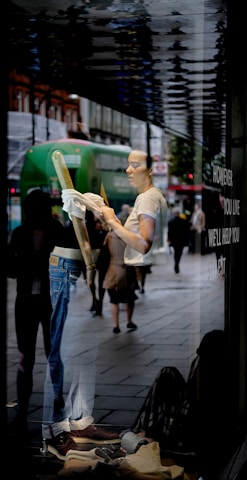 A person is seen cleaning a shop window from the inside, with reflections of a busy street outside. The person is wearing casual clothes and is holding a cloth. The display features jeans and brown shoes. Pedestrians and a green bus are visible in the reflection.
