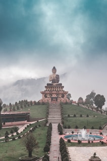 A large, serene Buddha statue is situated on an elevated platform in the center of a green, manicured landscape. A long staircase leads up to the statue, which is surrounded by well-maintained paths and trimmed bushes. In the foreground, there is a circular fountain with water jets creating a serene ambiance. The background features misty, cloud-covered hills or mountains, adding a mystical feel to the scene.