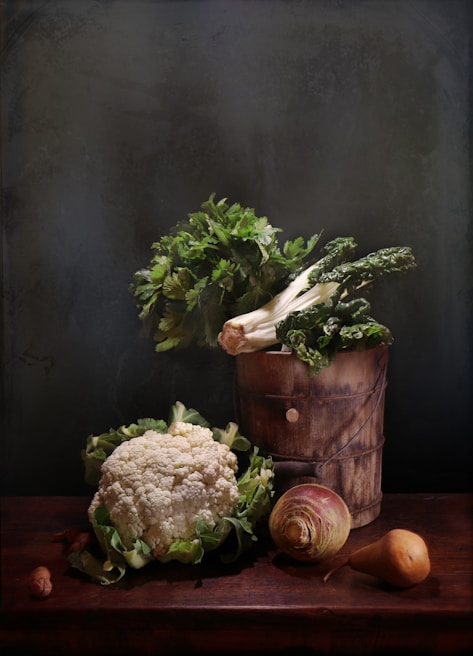 A rustic wooden table serves as the base for an arrangement of fresh vegetables. A bundle of leafy greens protrudes from a wooden bucket, surrounded by a cauliflower, a turnip, and a pear. The backdrop is a dark, textured wall that enhances the earthy tones and the natural appearance of the produce.