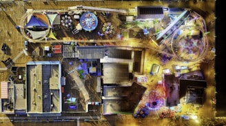 An aerial view of an amusement park with a variety of attractions including a roller coaster, a Ferris wheel, and other rides. The scene is brightly lit with colorful lights illuminating the area. There are tables with umbrellas and structures with multicolored roofs scattered throughout the park. Pathways weave among the attractions, and shadows are visible, suggesting a light source from above.