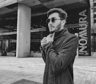 A stylish man adjusting his modern jacket collar while standing against a sleek urban wall.
