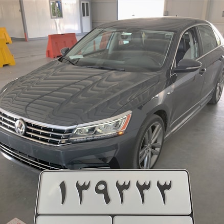 A dark gray sedan with a sporty design, parked inside a spacious warehouse-like garage with a concrete floor and metal walls. The front grille features a prominent logo, and the car's sleek headlights are visible. In the foreground, a detachable license plate with large, bold numerals in an unfamiliar script is shown. Brightly colored traffic barriers are positioned in the background.