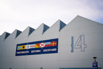 A large white industrial building with a series of peaked roofs features prominently. A colorful sign with nautical flags adorns the side, accompanied by the text 'PORTSMOUTH HISTORIC DOCKYARD' and 'Boathouse 4'. A small sign in the foreground reads 'Harbour Tours Waterbus'.
