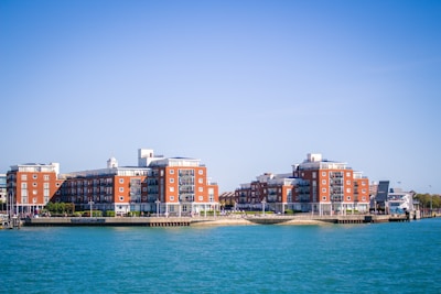 Two large red-brick residential buildings with balconies are situated by the water. The complex is surrounded by greenery and a few smaller structures can be seen in the vicinity. The sky is clear and blue, reflecting on the calm water of the river.