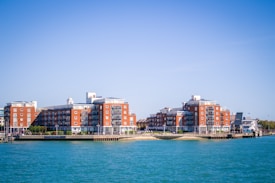 Two large red-brick residential buildings with balconies are situated by the water. The complex is surrounded by greenery and a few smaller structures can be seen in the vicinity. The sky is clear and blue, reflecting on the calm water of the river.