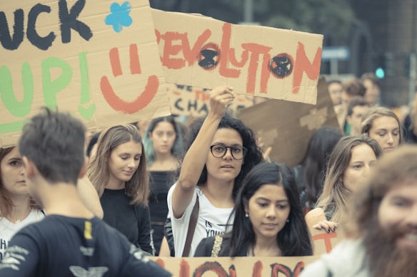 A large group of people is gathered, holding various cardboard signs. The signs have messages related to activism or protest, with visible text like 'REVOLUTION' and smiley faces. The crowd consists of diverse individuals, mostly young adults, who appear engaged and focused.