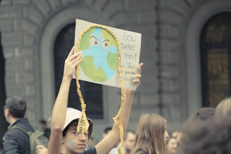 A protestor holding a sign with a depiction of Earth and an expression of concern. The sign includes text criticizing the treatment of the planet. Several people are visible in the background, suggesting a group gathering or protest.