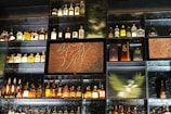 A wall of shelves filled with various bottles of liquor, including whiskey, gin, and other spirits. The shelves are arranged in a grid pattern and feature textured glass and decorative panels with earthy and abstract designs. Soft lighting highlights the bottles, creating an inviting and sophisticated atmosphere.