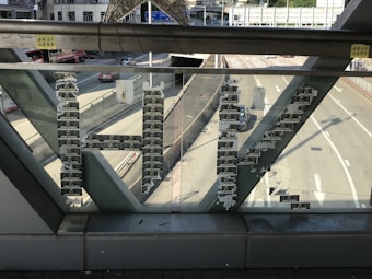 Stickers form the letters 'HK' on the glass of a pedestrian bridge, with a road and buildings visible in the background.