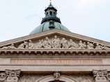 A classical architectural structure with ornate sculptures along the pediment. The sculptures depict various figures in seated and reclining positions, carved in a detailed, traditional style. The text 'EGO SUM VIA VERITAS ET VITA' is prominently displayed beneath the sculptures in gold lettering.