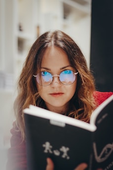 A woman with long brown hair is wearing round glasses with blue reflective lenses. She appears to be reading a book with a dark cover featuring white text and designs. The setting suggests an indoor environment with soft lighting, possibly a library or a study area.