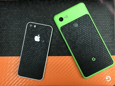 Two smartphones are placed on a textured surface. One phone has a green frame with a black honeycomb-patterned back and a fingerprint sensor. The other phone has a black and white color scheme with a similar honeycomb pattern and a prominent logo.