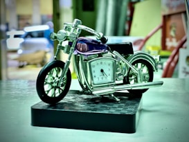 A miniature motorcycle model integrates a small clock where the engine would typically be. The model is placed on a patterned black base, and the surroundings are slightly blurred, suggesting an indoor location. Chrome elements and a transparent, dark purple piece adorn the motorcycle.