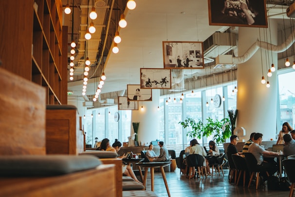 A modern cafe with a spacious and cozy interior is filled with people sitting at wooden tables and chairs. Soft lighting from numerous hanging bulbs creates a warm ambiance. Large windows allow natural light to flood in, and several framed photographs hang from the ceiling, adding an artistic touch. Potted plants are placed strategically around the room, enhancing the relaxed atmosphere.