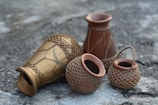A group of earthen pots arranged on a rough, stone-like surface. The pots vary in shape and size, featuring intricate net-like patterns around them. The texture and design suggest craftsmanship and a rustic aesthetic.