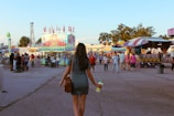 A young woman in a striped dress walks towards a brightly lit carnival area. Numerous people are visible, milling around various colorful stalls and attractions. There's a 'Sugar Barn' stand selling fair treats like apples and popcorn. The scene is lively with a mix of attendees, and the sky is clear with a few clouds.