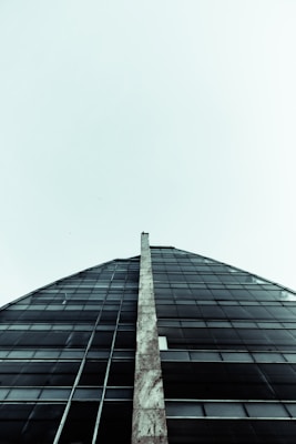 An upward perspective of a modern building with a glass facade. A tall, narrow, stone pillar is located in the center, reaching towards the sky. The structure has a triangular shape, emphasizing its height and sleek design.