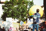 A crowd of people is gathered for a protest or demonstration, with someone holding a megaphone facing the group. Various signs with messages advocating for environmental action are visible. A large yellow balloon with a sun symbol floats above the crowd.