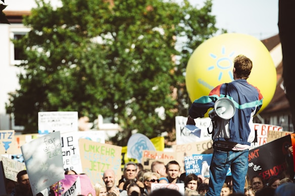 A crowd of people is gathered for a protest or demonstration, with someone holding a megaphone facing the group. Various signs with messages advocating for environmental action are visible. A large yellow balloon with a sun symbol floats above the crowd.