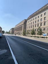 A wide street lined with a large, historic-looking government building on the right. The building features classical architecture with columns and a beige stone facade. There are trees planted along the sidewalk, and a few vehicles are parked on the left. The sky is clear and blue, suggesting a sunny day.