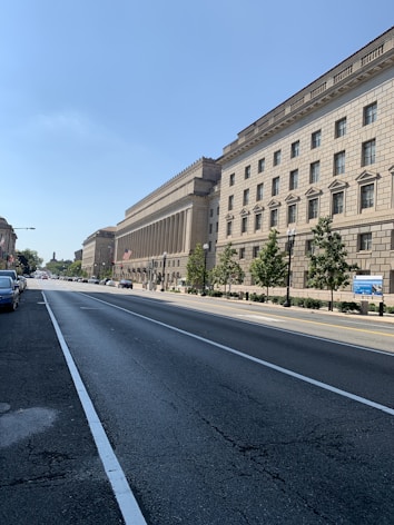 A wide street lined with a large, historic-looking government building on the right. The building features classical architecture with columns and a beige stone facade. There are trees planted along the sidewalk, and a few vehicles are parked on the left. The sky is clear and blue, suggesting a sunny day.