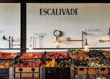 Wooden crates filled with various vegetables such as tomatoes, peppers, eggplants, and squash are displayed on a rustic wooden counter in a visually pleasing manner. Industrial-style light fixtures hang above, along with a decorative clock. The wall features artistic text labels in French indicating different types of produce.
