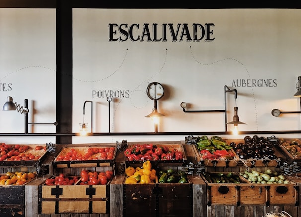 Wooden crates filled with various vegetables such as tomatoes, peppers, eggplants, and squash are displayed on a rustic wooden counter in a visually pleasing manner. Industrial-style light fixtures hang above, along with a decorative clock. The wall features artistic text labels in French indicating different types of produce.