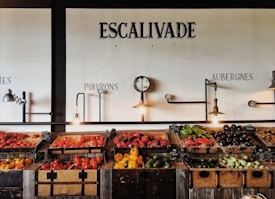 Wooden crates filled with various vegetables such as tomatoes, peppers, eggplants, and squash are displayed on a rustic wooden counter in a visually pleasing manner. Industrial-style light fixtures hang above, along with a decorative clock. The wall features artistic text labels in French indicating different types of produce.