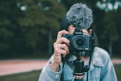 A person holding a Canon EOS camera equipped with a fluffy microphone, aimed directly at the viewer. The background shows an outdoor setting with out-of-focus greenery. The individual is partially visible, wearing a light denim jacket.
