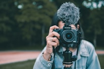 A person holding a Canon EOS camera equipped with a fluffy microphone, aimed directly at the viewer. The background shows an outdoor setting with out-of-focus greenery. The individual is partially visible, wearing a light denim jacket.