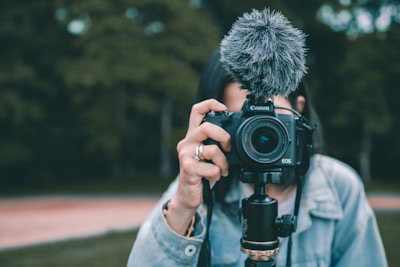 A person holding a Canon EOS camera equipped with a fluffy microphone, aimed directly at the viewer. The background shows an outdoor setting with out-of-focus greenery. The individual is partially visible, wearing a light denim jacket.