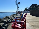 A row of brightly colored Adirondack chairs in red, blue, and yellow are lined up along a wooden boardwalk, with a backdrop of a waterfront and rocks. The word 'CANADA' is visible in large letters. Street lamps are positioned along the boardwalk, and there are buildings and cranes in the background.