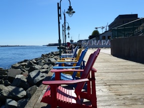A row of brightly colored Adirondack chairs in red, blue, and yellow are lined up along a wooden boardwalk, with a backdrop of a waterfront and rocks. The word 'CANADA' is visible in large letters. Street lamps are positioned along the boardwalk, and there are buildings and cranes in the background.