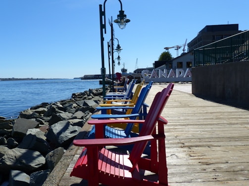 A row of brightly colored Adirondack chairs in red, blue, and yellow are lined up along a wooden boardwalk, with a backdrop of a waterfront and rocks. The word 'CANADA' is visible in large letters. Street lamps are positioned along the boardwalk, and there are buildings and cranes in the background.