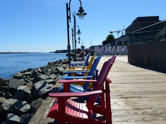 A row of brightly colored Adirondack chairs in red, blue, and yellow are lined up along a wooden boardwalk, with a backdrop of a waterfront and rocks. The word 'CANADA' is visible in large letters. Street lamps are positioned along the boardwalk, and there are buildings and cranes in the background.