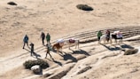 A group of people walking with pack animals on a rugged, dirt trail in an arid landscape. There are five individuals dressed in outdoor gear and three pack animals, each carrying loads. The path and surrounding area are dry and dusty with sparse vegetation and some bushes.