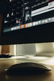 A sleek, white computer mouse rests on a smooth surface, partially illuminated by the screen of a large desktop monitor. The monitor displays a dimly lit interface with various icons and tools, suggesting image or video editing software.