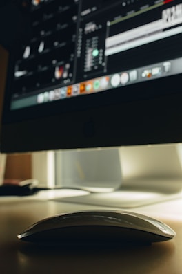 A sleek, white computer mouse rests on a smooth surface, partially illuminated by the screen of a large desktop monitor. The monitor displays a dimly lit interface with various icons and tools, suggesting image or video editing software.