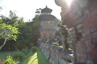 A traditional structure in a lush green setting is illuminated by sunlight filtering through trees. The building features brickwork and intricate carvings, surrounded by well-maintained gardens.