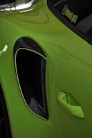 A close-up of a bright green car showing the area around the side air intake and door handle. The car's paint reflects light, and a window decal with text is visible on the edge of the window.