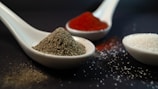Ceramic spoons filled with different spices and ingredients are arranged on a dark surface. One spoon contains coarse black pepper, another holds bright red paprika, and a third is filled with crystalline white sugar. Small granules are scattered around, enhancing the focus on the textures of the spices.
